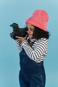 Curious black girl with curly hair in stylish clothes taking photo on instant retro photo camera against blue background in studio